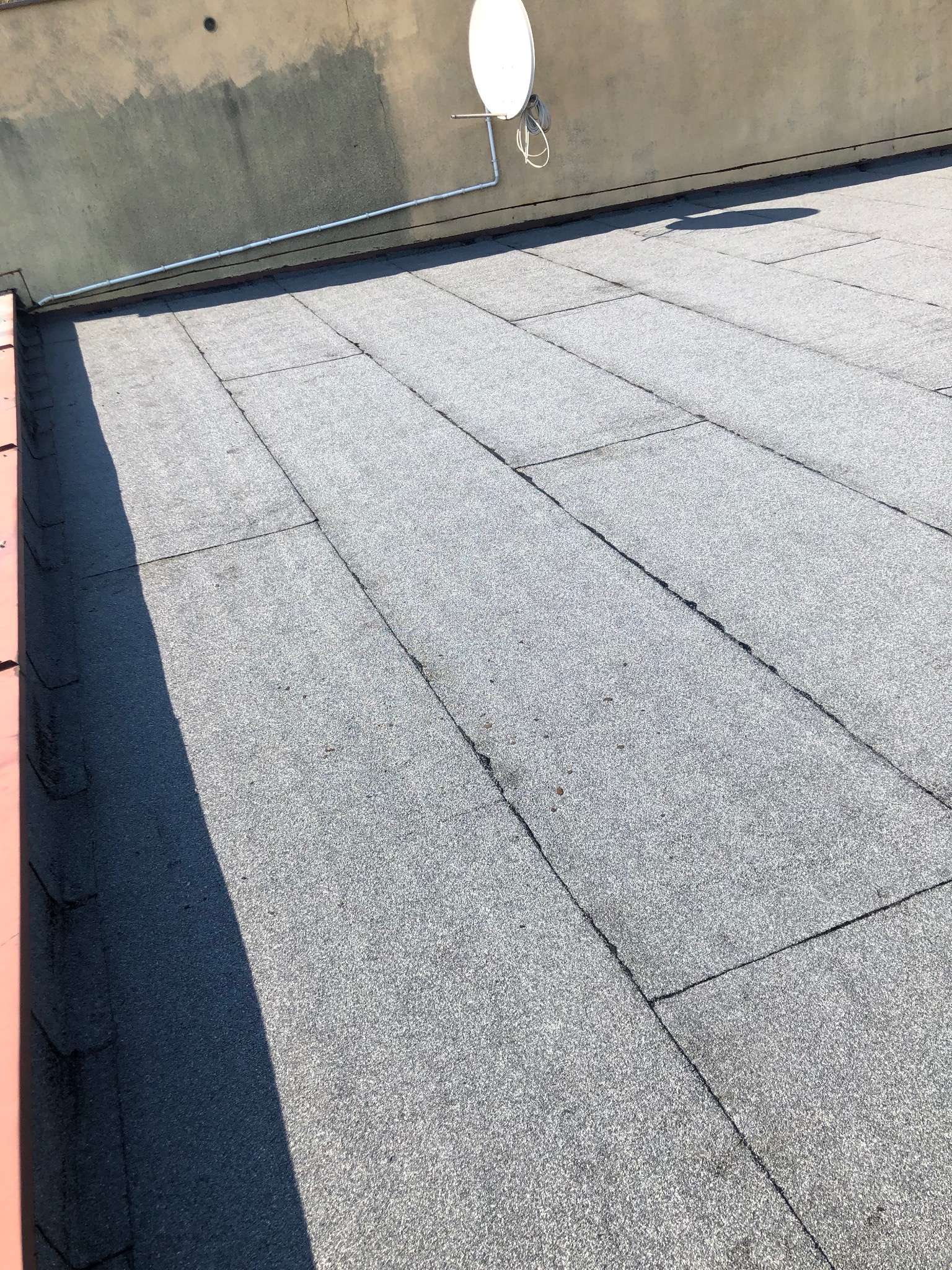 Szary dach pokryty papą bitumiczną w prostokątnych panelach, z anteną satelitarną w tle i cieniem na lewym brzegu.