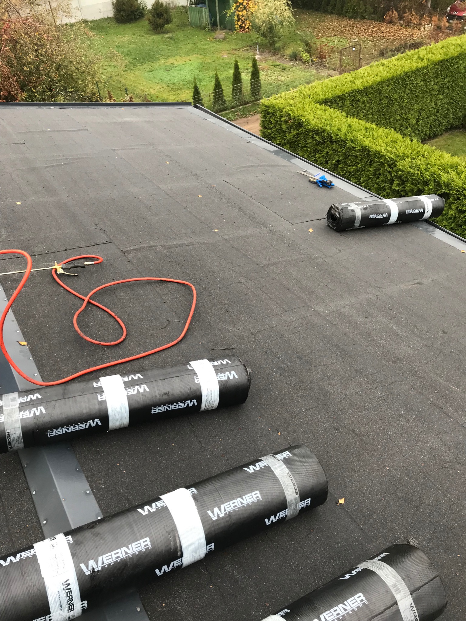 Widok z góry na płaski dach pokryty papą, z widocznymi rolkami papy marki Werner, narzędziami dekarskimi i zielonym otoczeniem w tle.