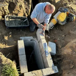 Pracownik w trakcie budowy betonowej studzienki w wykopie, używając poziomicy i gumowego młotka, obok ubijarka wibracyjna i pojemnik z zaprawą.