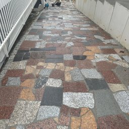 FIBRO SPÓŁKA Z OGRANICZONĄ ODPOWIEDZIALNOŚCIĄ - Szeroki chodnik wyłożony nieregularnymi płytami z kamienia w odcieniach szarości, brązu i czerwieni, z pracownikiem w tle przygotowującym zaprawę, obok biała balustrada i betonowe zabezpieczenie...
