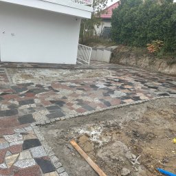 FIBRO SPÓŁKA Z OGRANICZONĄ ODPOWIEDZIALNOŚCIĄ - Ułożona kolorowa kostka granitowa przed nowoczesnym domem z widocznym fragmentem terenu przygotowywanego pod dalsze prace brukarskie oraz niebieski przewód instalacyjny