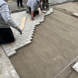 FIBRO SPÓŁKA Z OGRANICZONĄ ODPOWIEDZIALNOŚCIĄ - Trzech mężczyzn w trakcie układania szarej kostki brukowej na przygotowanym podłożu z piasku. Widoczne narzędzia i fragmenty już ułożonej nawierzchni.