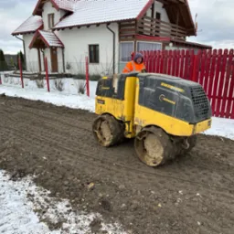 Operator w pomarańczowej kamizelce obsługuje żółty walec drogowy Bomag BMP8500III na placu budowy z częściowo stopniałym śniegiem, na tle białego domu z czerwonym dachem i czerwonego drewnianego...
