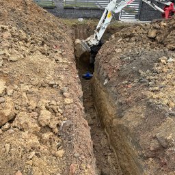 FIBRO SPÓŁKA Z OGRANICZONĄ ODPOWIEDZIALNOŚCIĄ - Wąski wykop w ziemi, wewnątrz widoczny niebieski zawór, nad nim ramię koparki w trakcie pracy, obok hałda ziemi, w tle ogrodzenie posesji.