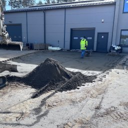 Fibro Budownictwo - Układanie kostki brukowej na placu: koparka, pracownik zagęszcza podłoże, widoczny fragment obrzeża z kostki. Praca w toku, przygotowanie podłoża pod kostkę.