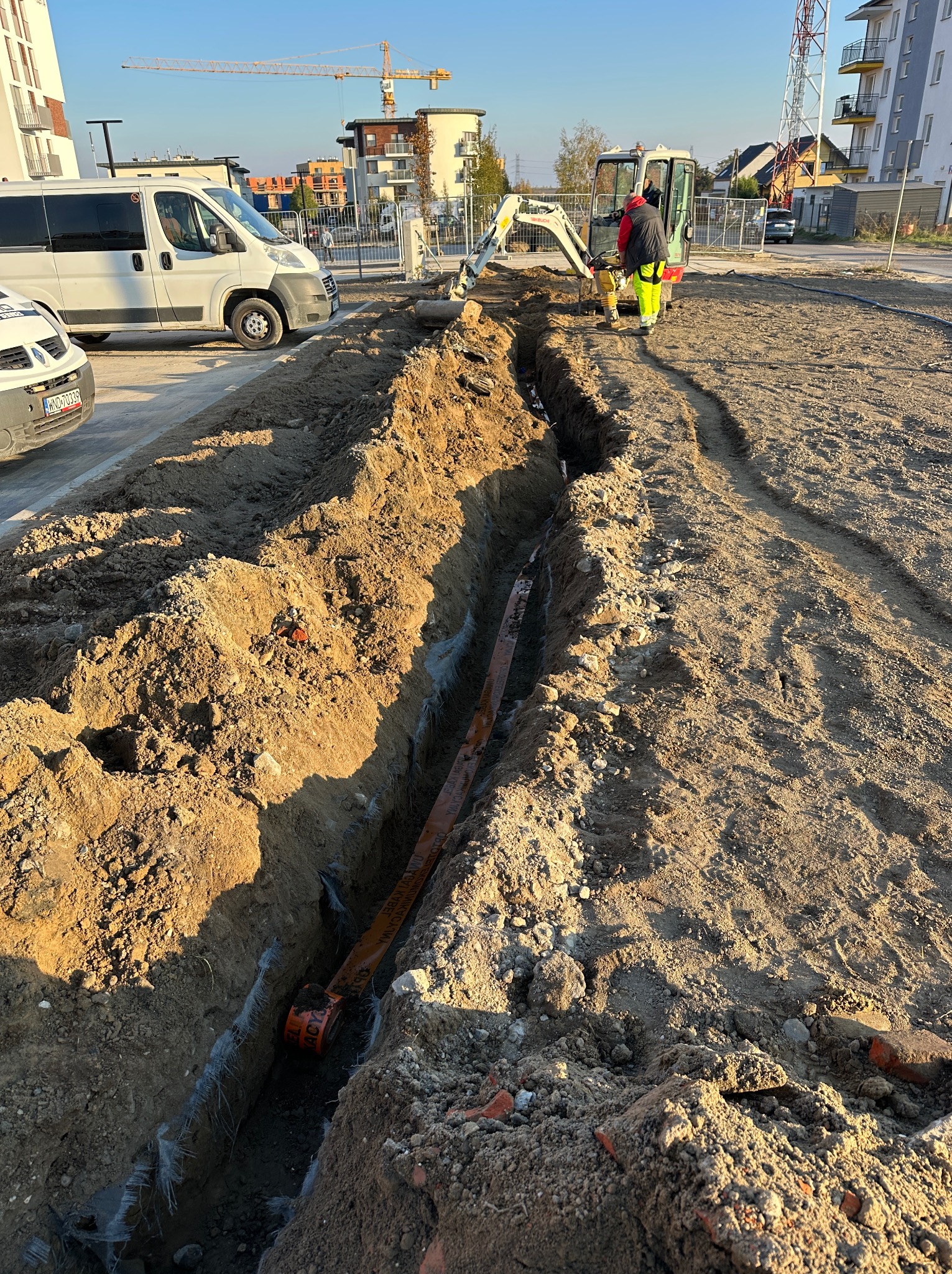 Wykop liniowy w ziemi z widoczną pomarańczową rurą wewnątrz, mała koparka i pracownik w odblaskowej kamizelce w tle, teren budowy z nowymi budynkami i pojazdami w tle.