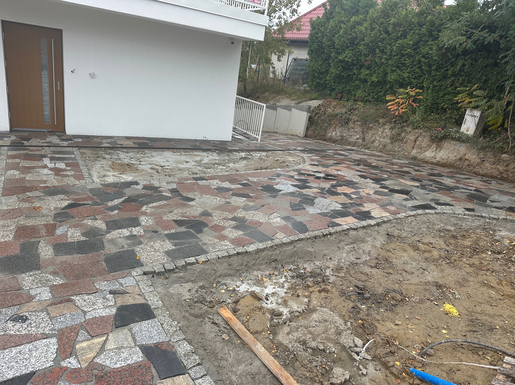 Ułożona kolorowa kostka granitowa przed nowoczesnym domem z widocznym fragmentem terenu przygotowywanego pod dalsze prace brukarskie oraz niebieski przewód instalacyjny