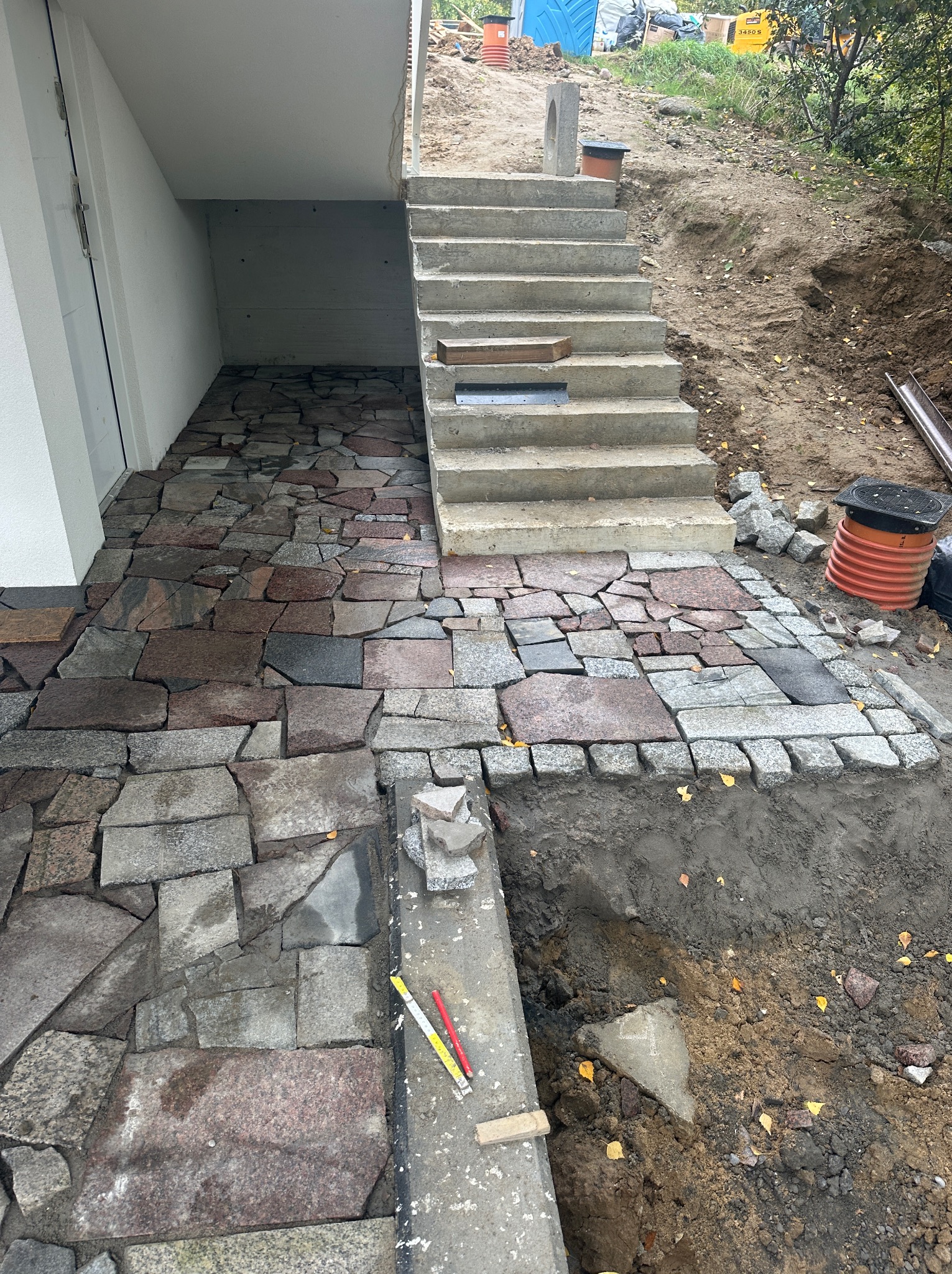 Fragment podwórza z nieregularną kamienną nawierzchnią w odcieniach szarości i czerwieni, przylegającą do betonowych schodów i wejścia do piwnicy, z widocznym wykopem i narzędziami pomiarowymi.