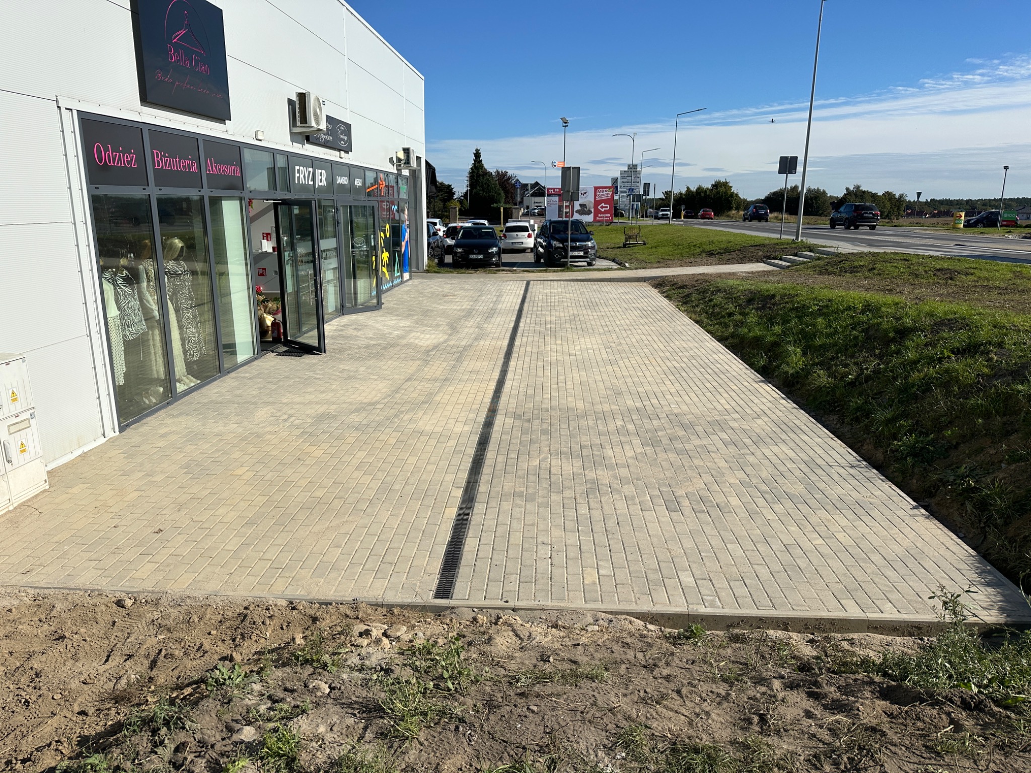 Nowo ułożona, jasna kostka brukowa przed budynkiem komercyjnym z widoczną kratką odpływową liniową pośrodku, teren wyrównany, przygotowany pod nasadzenia.