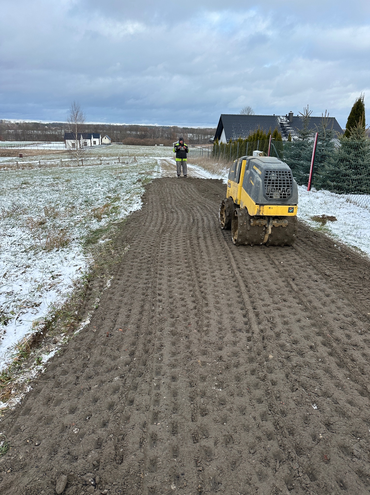 Żółto-czarny walec wibracyjny BOMAG ubija ziemię na działce w zimowy dzień, pracownik kontroluje postęp prac, w tle zabudowa jednorodzinna i resztki śniegu.
