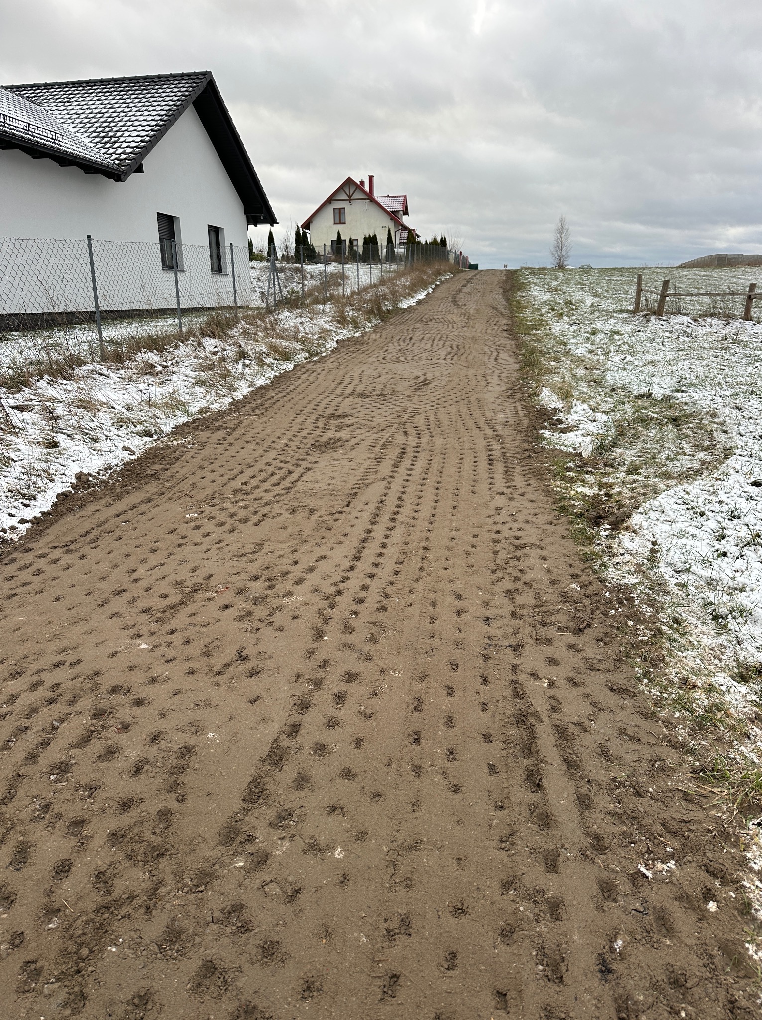Grząska, gruntowa droga po przejeździe ciężkiego sprzętu, widoczne ślady kół, w tle domy jednorodzinne i częściowo ośnieżone pole w pochmurny dzień.
