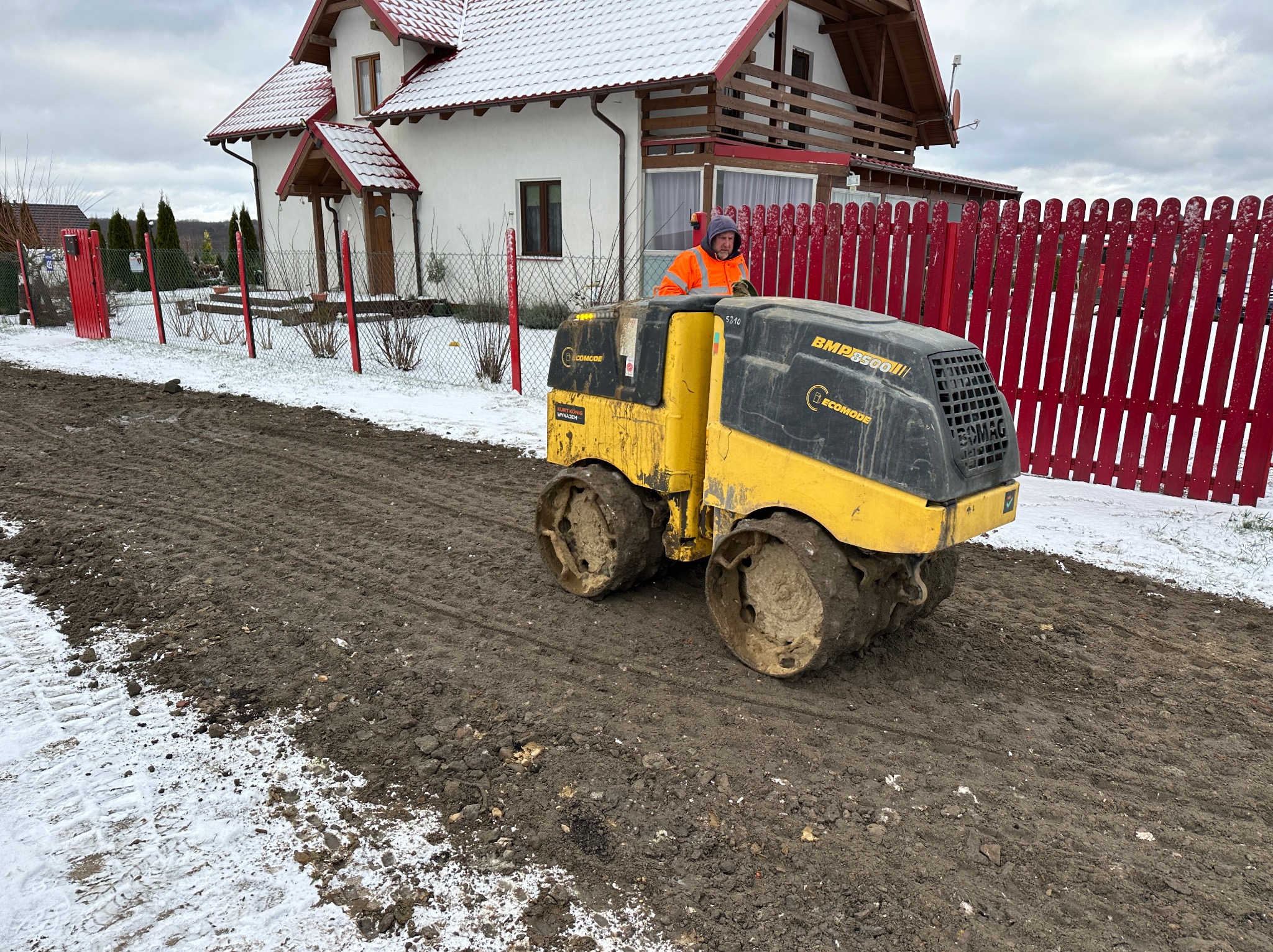 Operator w pomarańczowej kamizelce obsługuje żółty walec drogowy Bomag BMP8500III na placu budowy z częściowo stopniałym śniegiem, na tle białego domu z czerwonym dachem i czerwonego drewnianego...