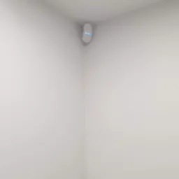 Biały czujnik alarmowy z niebieską diodą LED, zamontowany w rogu białego pokoju, widoczne przewody pod sufitem.