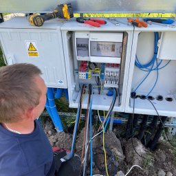 ES-COM SERVICE SPÓŁKA Z OGRANICZONĄ ODPOWIEDZIALNOŚCIĄ - Elektryk instaluje rozdzielnicę elektryczną na zewnątrz, widoczne narzędzia, kable w peszlach i tabliczka ostrzegawcza.