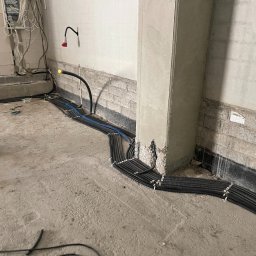 ES-COM SERVICE SPÓŁKA Z OGRANICZONĄ ODPOWIEDZIALNOŚCIĄ - Wiązka czarnych kabli w peszlach ułożona wzdłuż ściany w surowym wnętrzu, widoczna puszka elektryczna oraz wystające przewody.