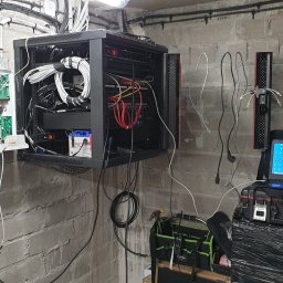 ES-COM SERVICE SPÓŁKA Z OGRANICZONĄ ODPOWIEDZIALNOŚCIĄ - Szafa rack z serwerami i okablowaniem zamontowana na ścianie z cegły, obok otwarta skrzynka z elektroniką, laptop na skrzynce narzędziowej, widoczne narzędzia i torba instalatora.