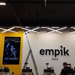 ES-COM SERVICE SPÓŁKA Z OGRANICZONĄ ODPOWIEDZIALNOŚCIĄ - Instalacja oświetlenia w sklepie Empik Aura w Olsztynie, widoczne krzyżowe lampy LED podwieszone pod sufitem, drabina rozstawiona, pracownik w czarnej kurtce przy kasie.