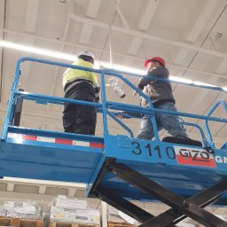 ES-COM SERVICE SPÓŁKA Z OGRANICZONĄ ODPOWIEDZIALNOŚCIĄ - Dwóch monterów na podnośniku koszowym instaluje kamerę przemysłową pod sufitem w hali magazynowej, widoczne rury instalacyjne pod stropem.