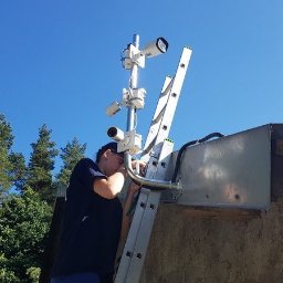 ES-COM SERVICE SPÓŁKA Z OGRANICZONĄ ODPOWIEDZIALNOŚCIĄ - Pracownik na drabinie instaluje system monitoringu z trzema kamerami na zewnątrz budynku, widok na niebieskie niebo i drzewa w tle.