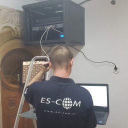 ES-COM SERVICE SPÓŁKA Z OGRANICZONĄ ODPOWIEDZIALNOŚCIĄ - Instalator w koszulce firmowej ES-COM konfiguruje szafę rack z urządzeniami sieciowymi na drabinie, z laptopem w ręku.