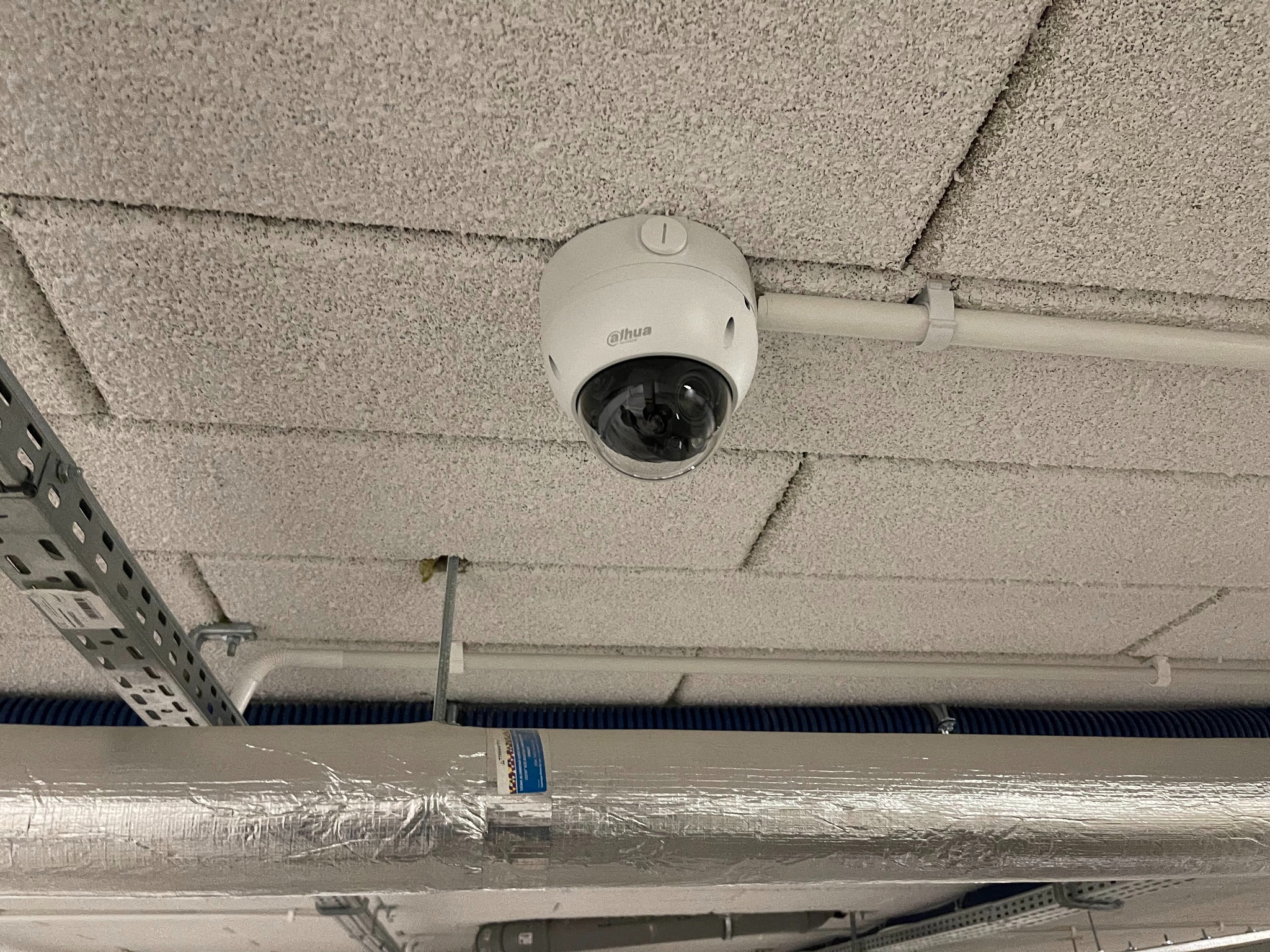 Kopułowa kamera monitoringu Dahua zamontowana na suficie podwieszanym w pomieszczeniu przemysłowym, widoczne rury i kable w metalowych osłonach.