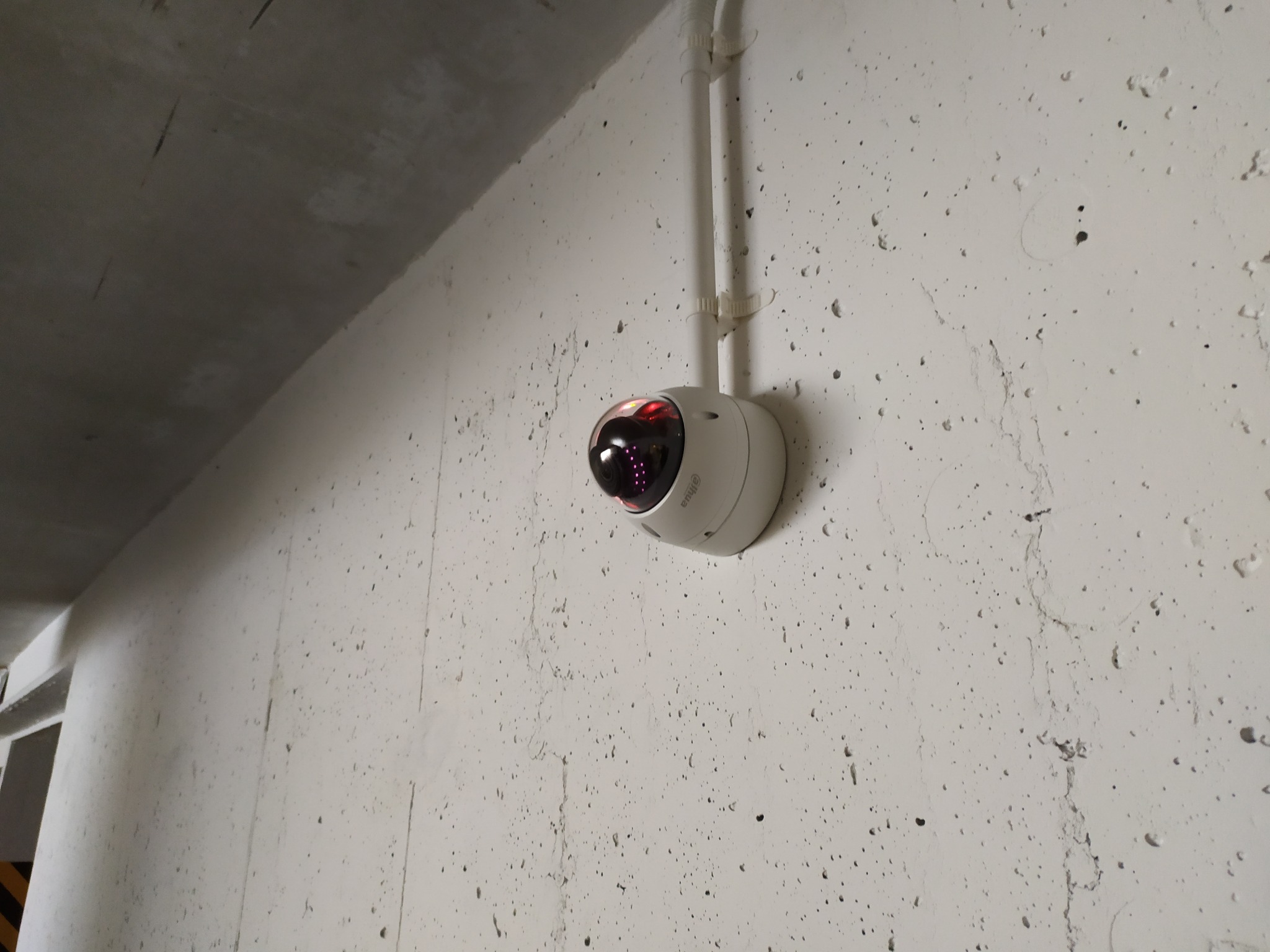 Kopułowa kamera monitoringu Dahua, zamontowana na szarej, porowatej ścianie w garażu podziemnym, z widocznymi przewodami w białej rurce PCV.