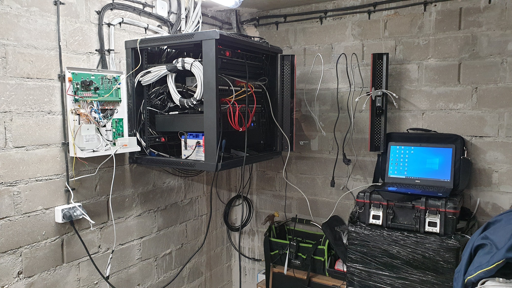 Szafa rack z serwerami i okablowaniem zamontowana na ścianie z cegły, obok otwarta skrzynka z elektroniką, laptop na skrzynce narzędziowej, widoczne narzędzia i torba instalatora.