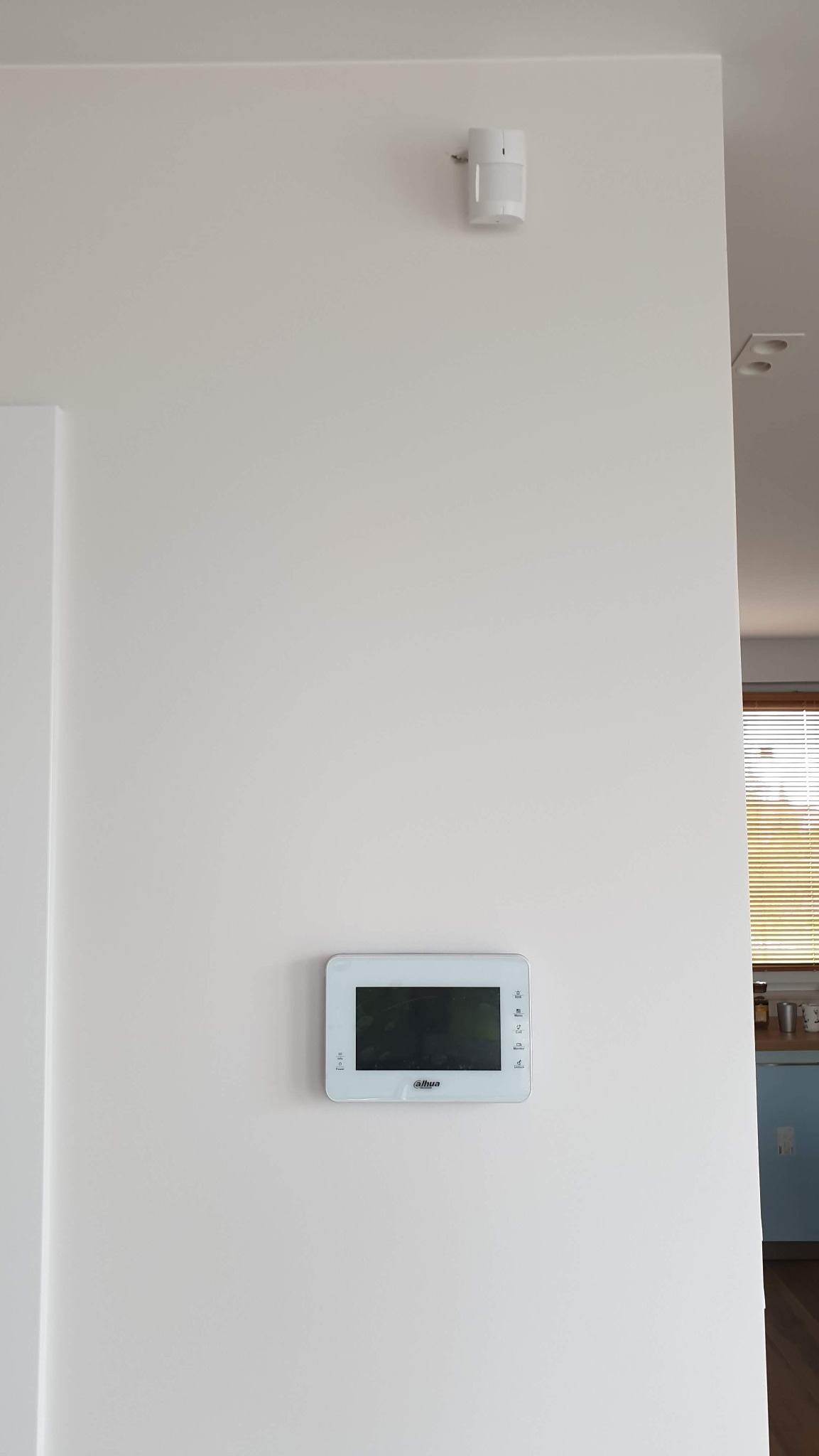 Biała ściana z zamontowanym panelem wideodomofonu marki Dahua i czujnikiem ruchu alarmu, widok na wnętrze przez otwarte drzwi.