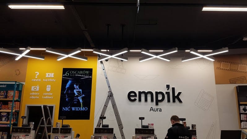 Instalacja oświetlenia w sklepie Empik Aura w Olsztynie, widoczne krzyżowe lampy LED podwieszone pod sufitem, drabina rozstawiona, pracownik w czarnej kurtce przy kasie.