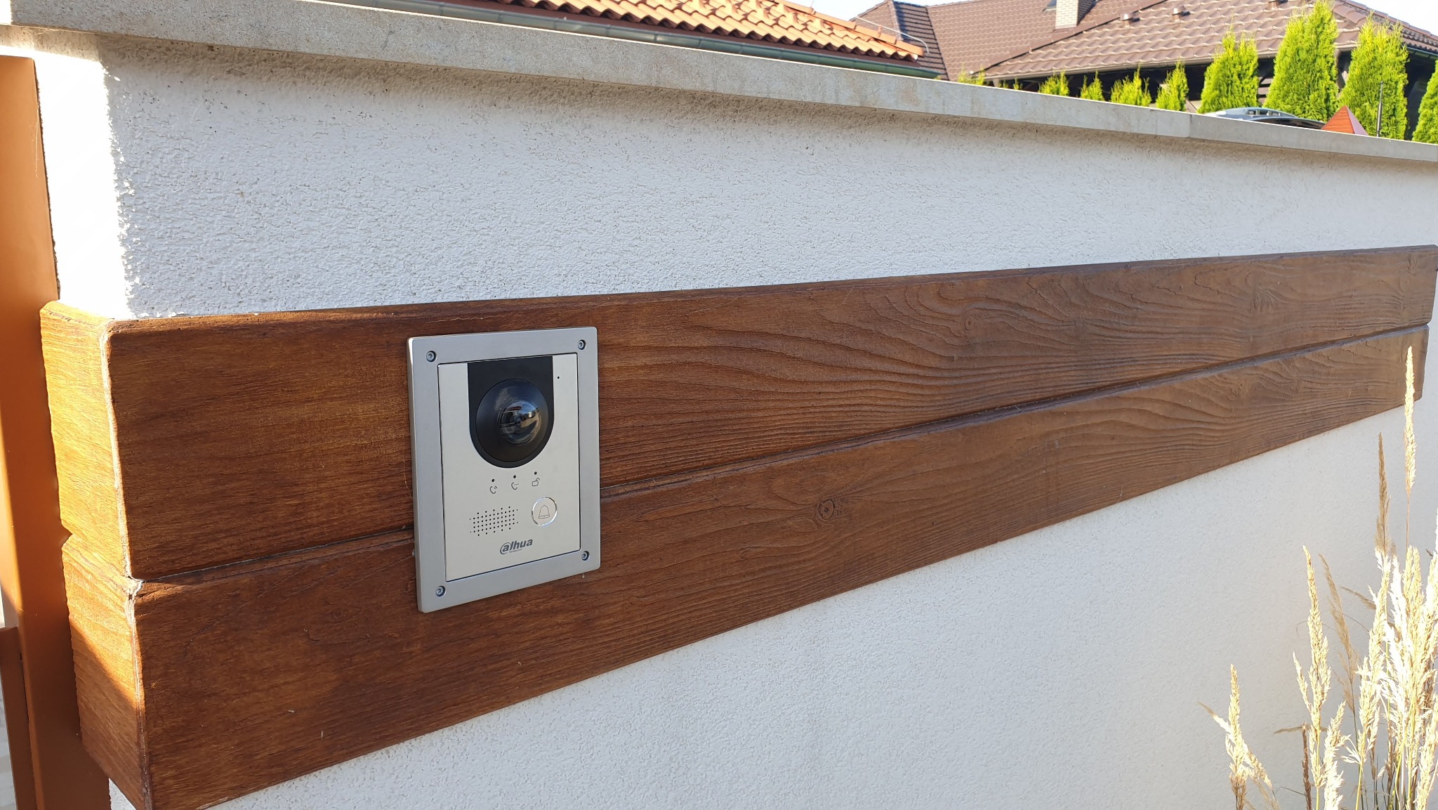 Zewnętrzny panel wideodomofonu Dahua zamontowany na drewnianym elemencie ogrodzenia z białym, teksturowanym tłem ściany i widocznymi elementami roślinności w tle.
