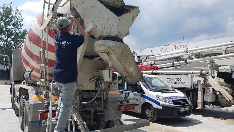 Pracownik na drabinie przy betonomieszarce z czerwono-białymi pasami, obok samochód z napisem 'Systemy Monitoringu' i pompa do betonu w tle.
