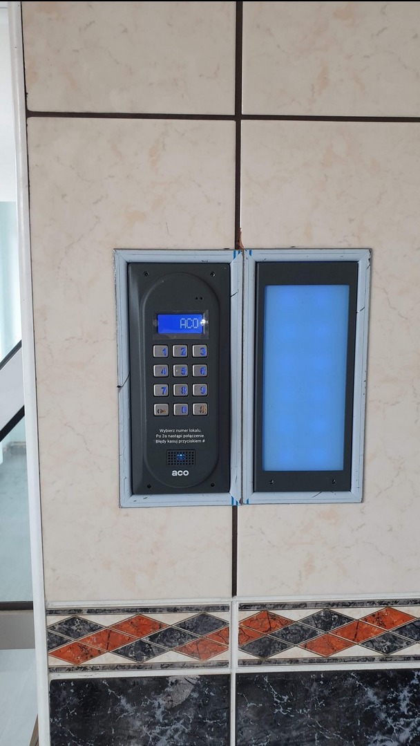 Panel domofonu z klawiaturą numeryczną i ekranem LCD, zamontowany na ścianie wyłożonej marmurkowymi płytkami z dekoracyjnym pasem. Widoczny napis 'ACO' na panelu.