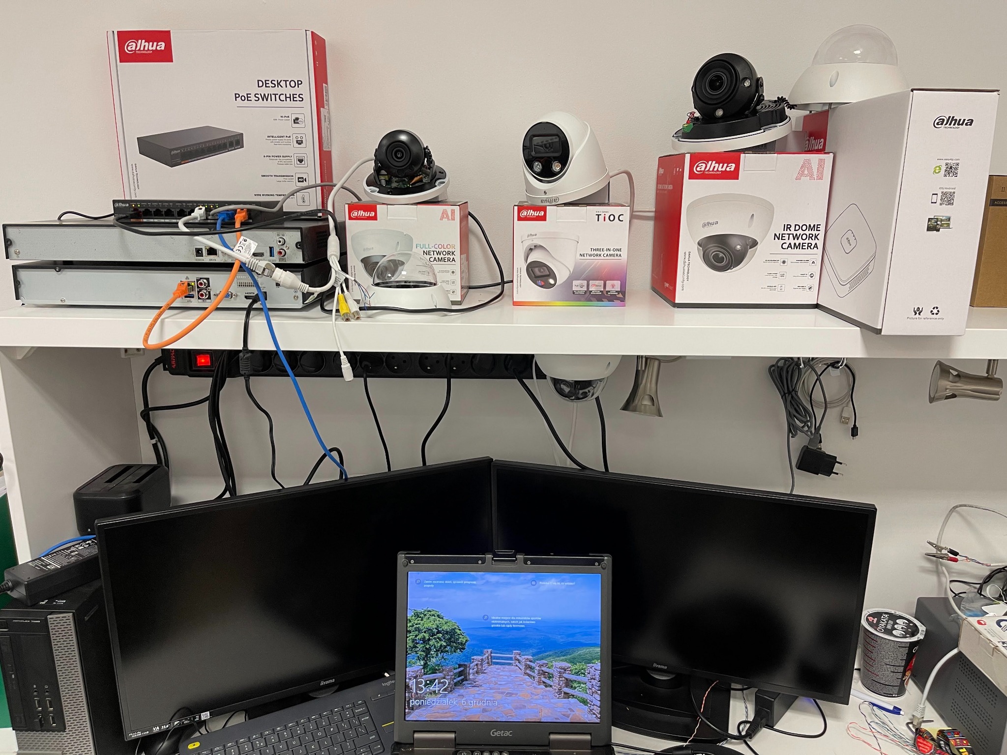 Półka z urządzeniami i pudełkami Dahua: kamery IP, rejestratory, przełączniki PoE. Na pierwszym planie dwa monitory i laptop Getac z widokiem na góry.