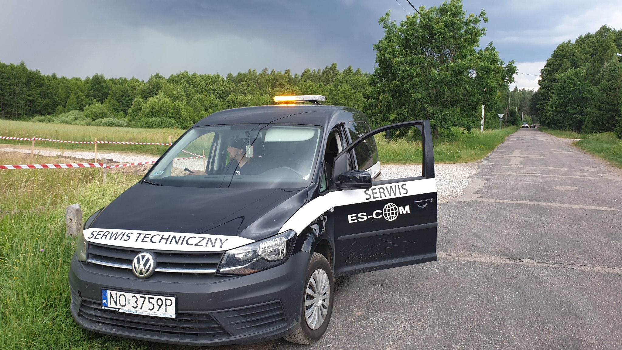 Czarny samochód serwisowy z otwartymi drzwiami i napisem 'SERWIS TECHNICZNY' na tle drogi i zalesionego terenu pod pochmurnym niebem.