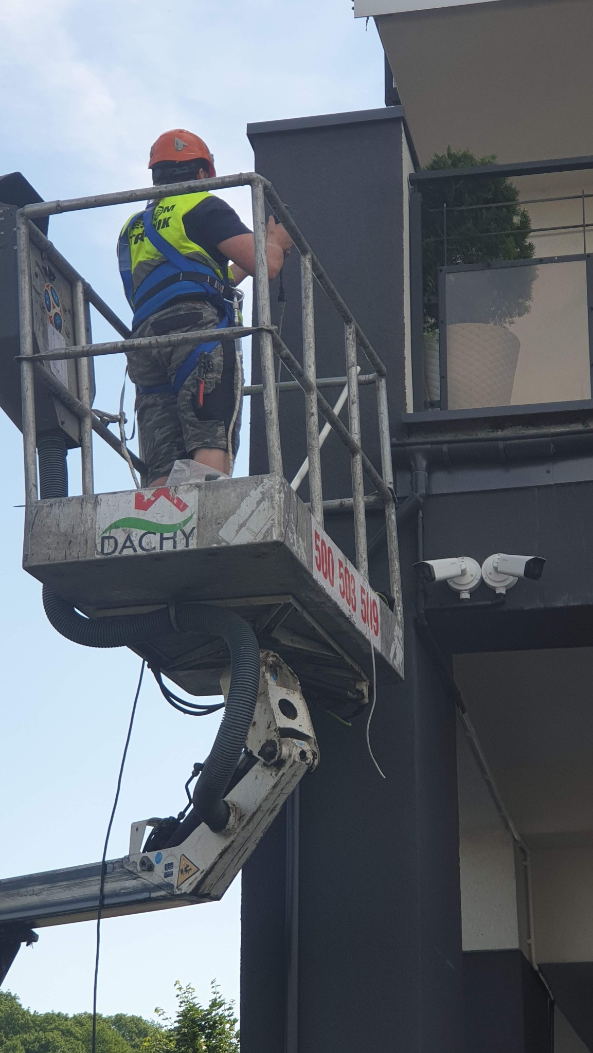 Pracownik w pomarańczowym kasku i żółtej kamizelce na podnośniku koszowym, montuje dwie białe kamery monitoringu na elewacji budynku.