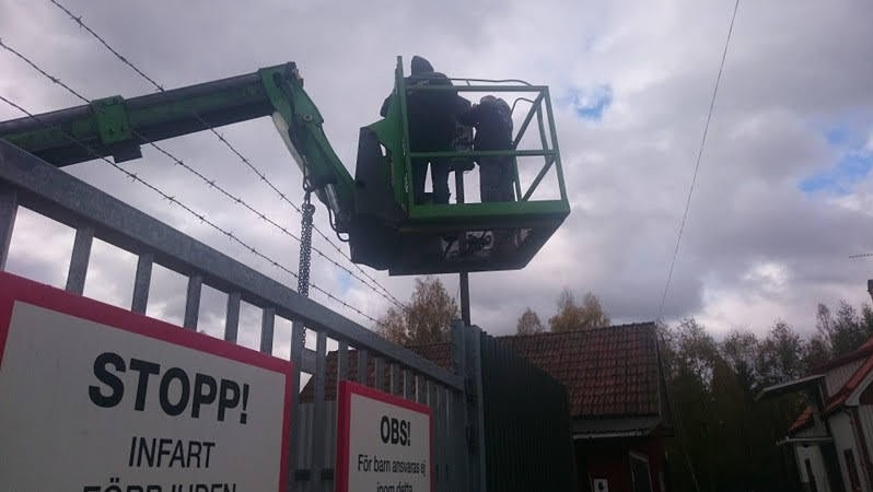Pracownicy w podnośniku koszowym instalują drut kolczasty na szczycie metalowej bramy z tablicami ostrzegawczymi w języku szwedzkim, widok z dołu na tle pochmurnego nieba.