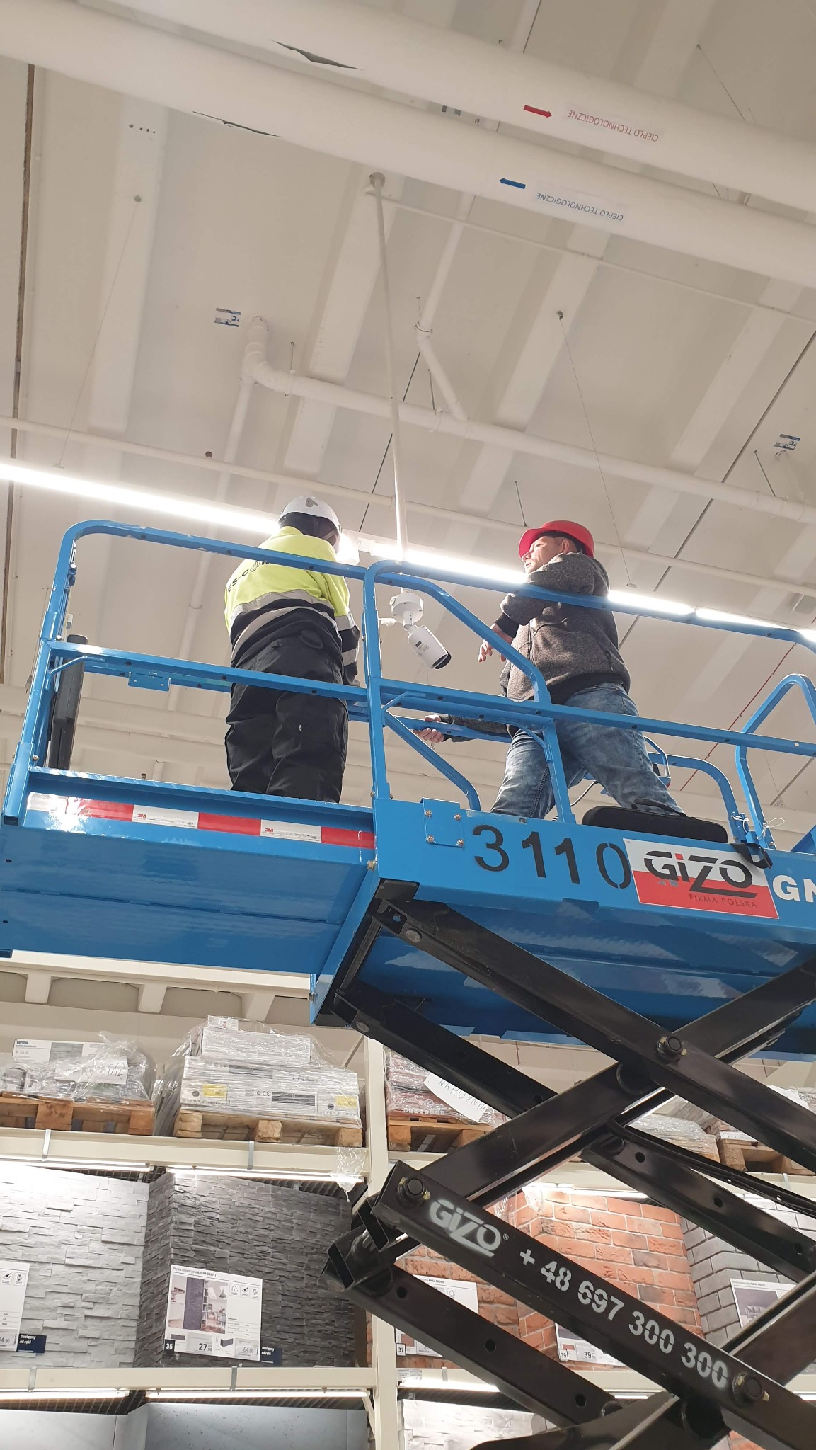 Dwóch monterów na podnośniku koszowym instaluje kamerę przemysłową pod sufitem w hali magazynowej, widoczne rury instalacyjne pod stropem.