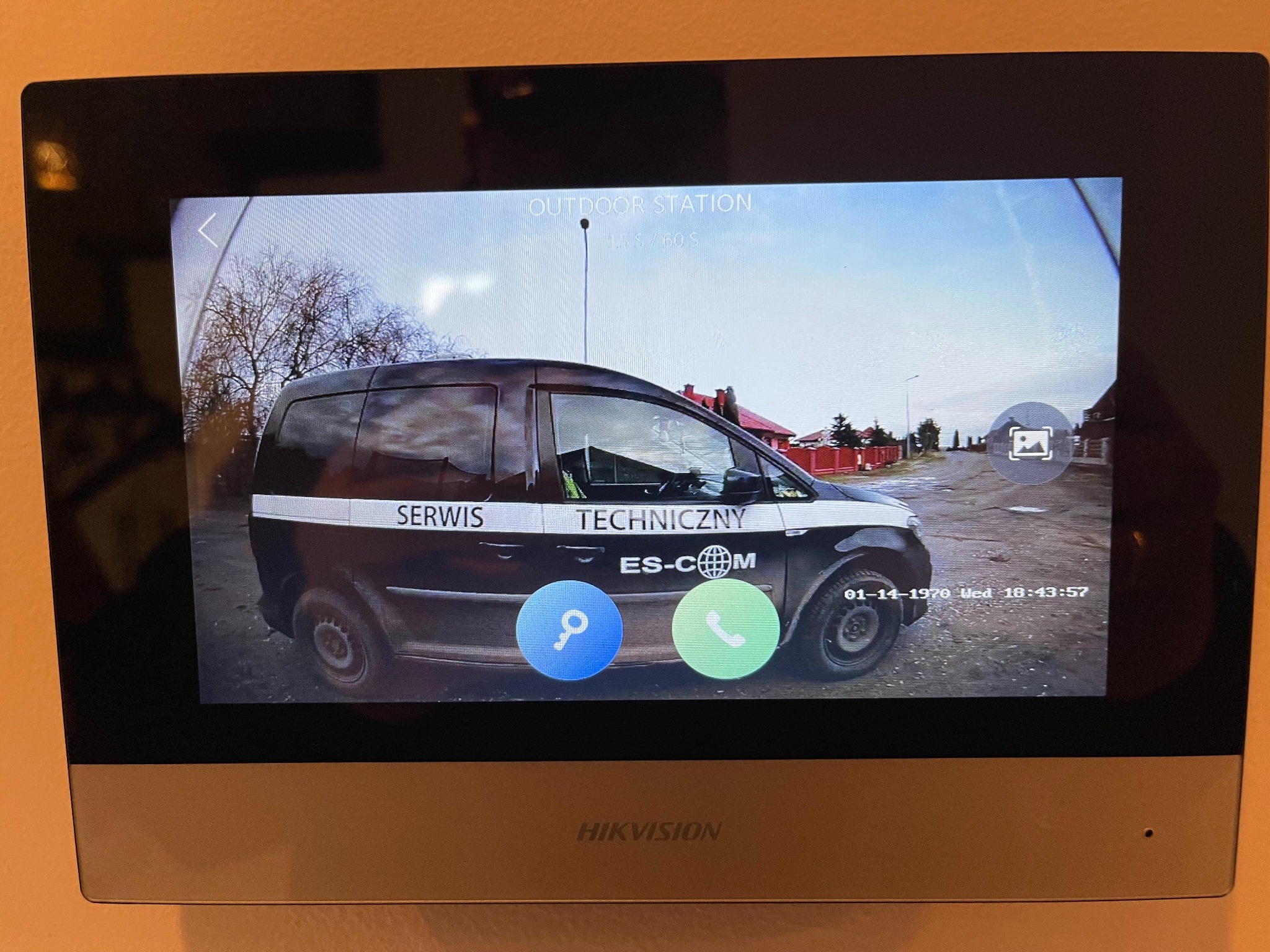 Ekran wideodomofonu Hikvision wyświetlający obraz z kamery: samochód serwisowy firmy ES-COM na tle ulicy i domów.