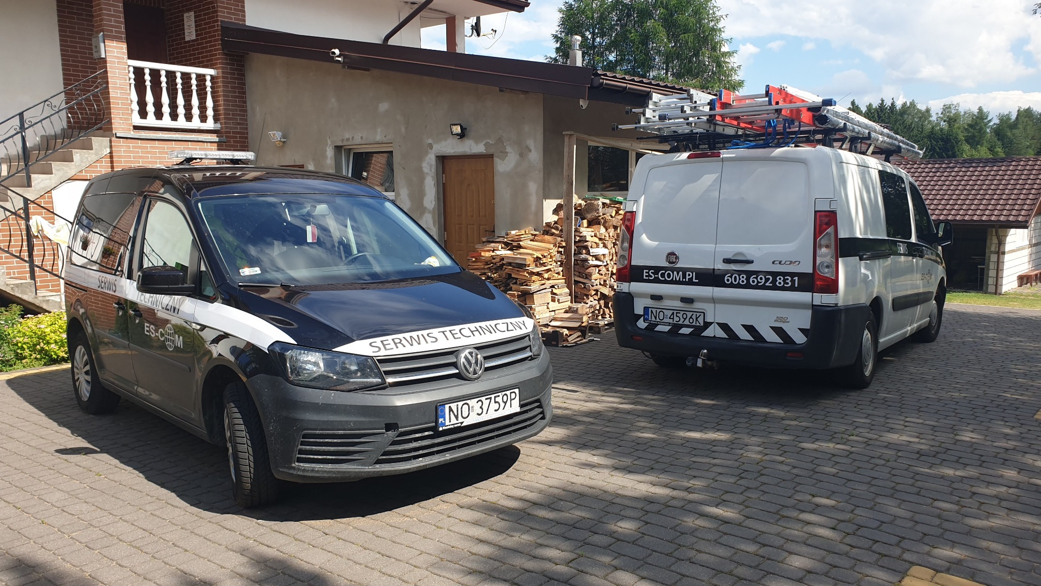 Dwa samochody serwisowe firmowe zaparkowane na brukowanym podjeździe przed domem, biały Fiat z drabiną na dachu i czarny Volkswagen z napisem 'Serwis Techniczny' na masce.