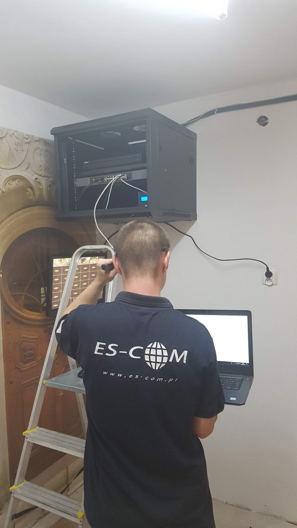 Instalator w koszulce firmowej ES-COM konfiguruje szafę rack z urządzeniami sieciowymi na drabinie, z laptopem w ręku.