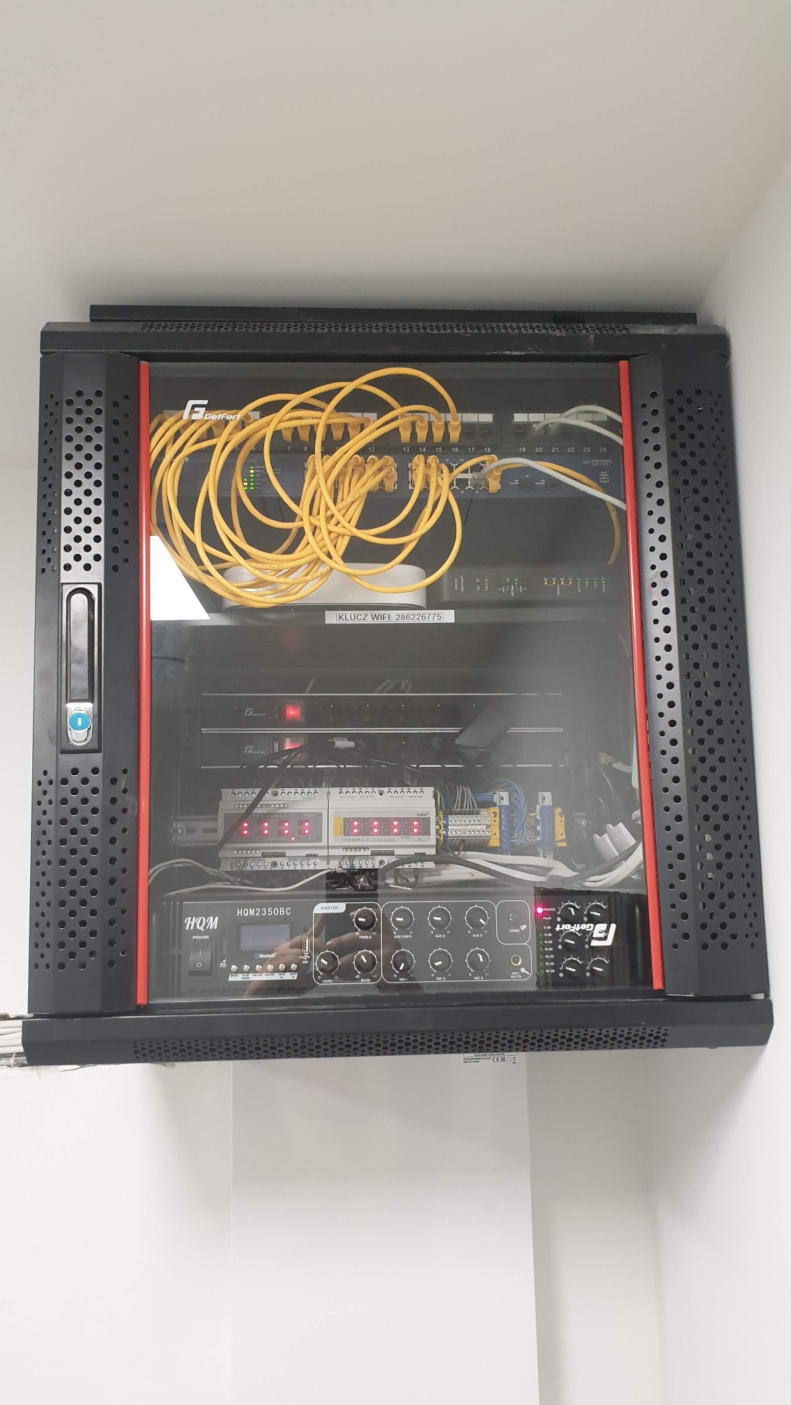 Zamknięta szafa rack z widocznymi kablami ethernet, routerem WiFi z naklejką 'KLUCZ WIFI: 286226775', modułami sterującymi i wzmacniaczem HQM2350BC oraz GetFort GS7241.