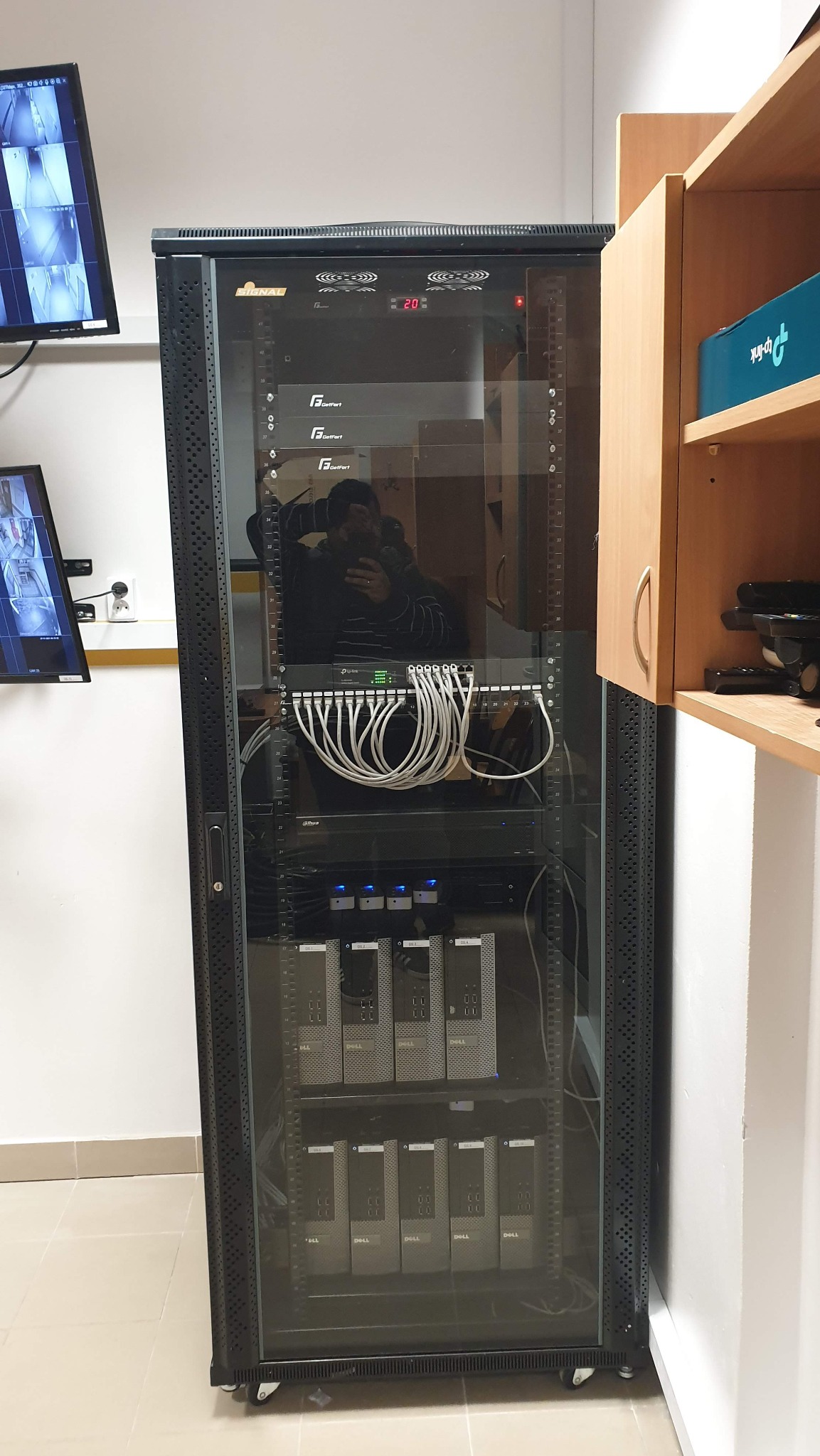 Szafa rack z zainstalowanymi urządzeniami, widoczne serwery Dell, switch TP-Link z podłączonymi kablami, na górze urządzenia marki GetFort, obok na ścianie monitory podglądu kamer.