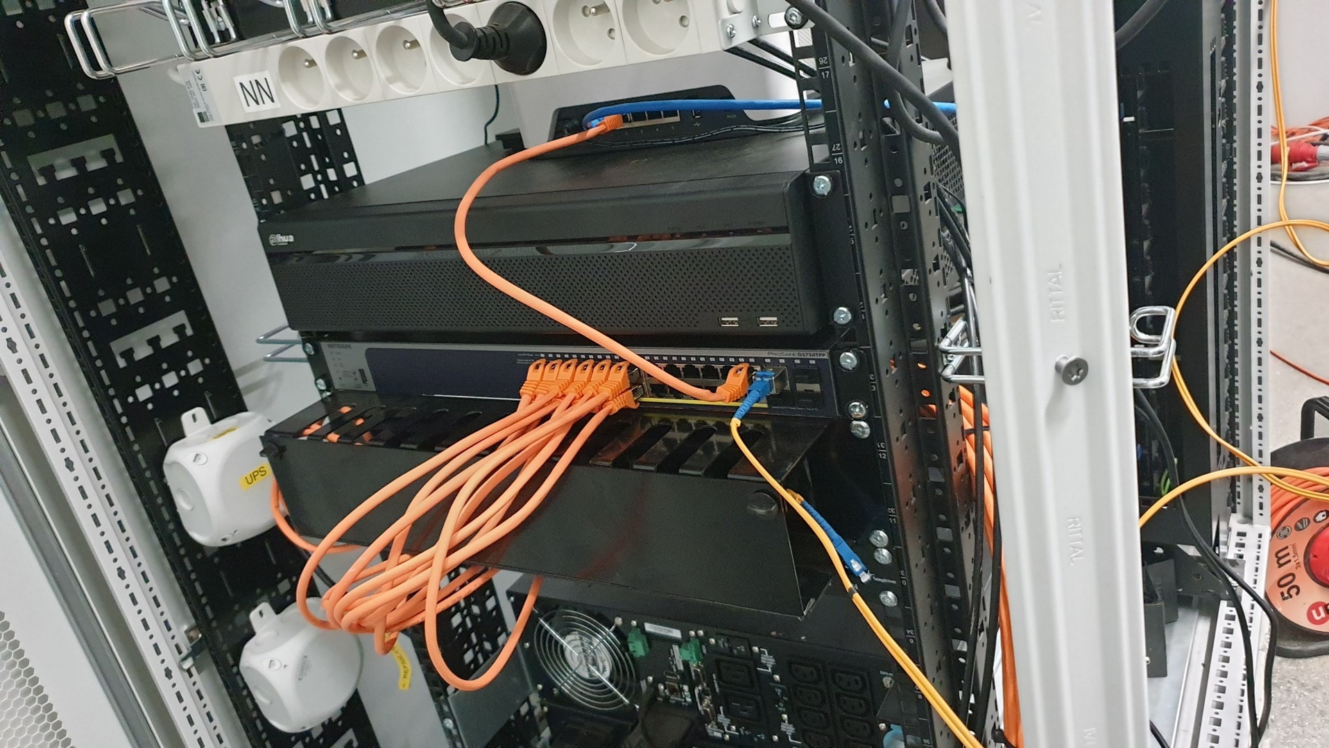 Serwerownia z zamontowanym rejestratorem Dahua, switchem Netgear i UPS-ami, pomarańczowe kable sieciowe podłączone do portów. Widoczna listwa zasilająca z wtyczkami europejskimi.