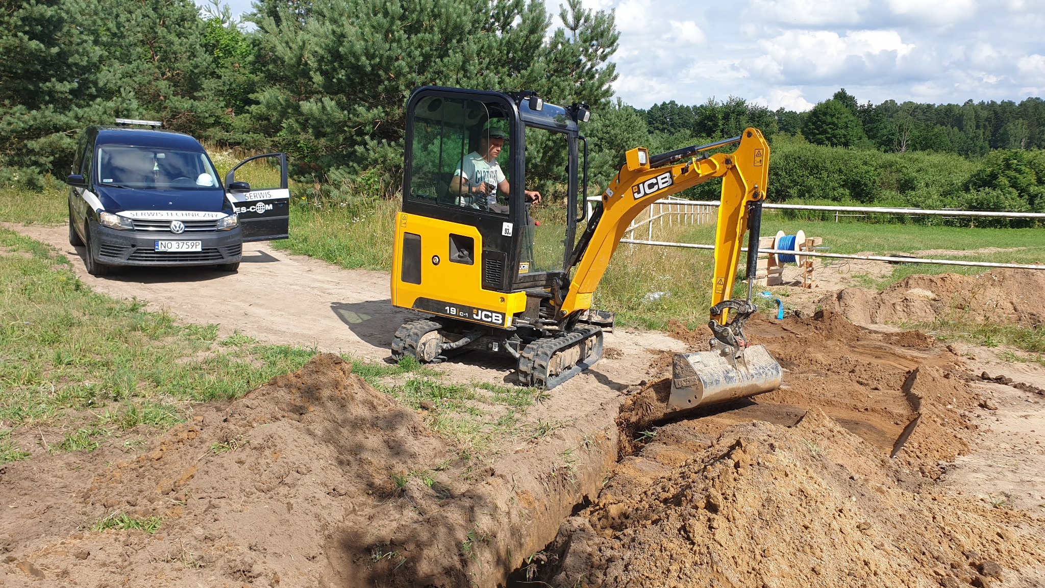 Żółta mini koparka JCB 19C-1 w trakcie kopania rowu w ziemi, obok zaparkowany samochód serwisowy z otwartymi drzwiami, w tle drzewa i niebo.