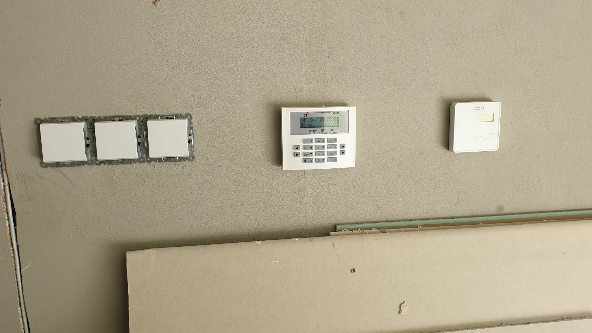 Centrala alarmowa z wyświetlaczem LCD i klawiaturą numeryczną zamontowana na szarej ścianie obok trzech gniazdek elektrycznych bez ramek, widoczny fragment płyty gipsowej.