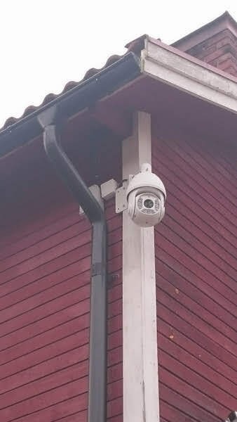 Kamera obrotowa PTZ zamontowana na białej kolumnie przy czerwonej drewnianej elewacji budynku, obok rynny. Widok z dołu, z pochmurnym niebem w tle.