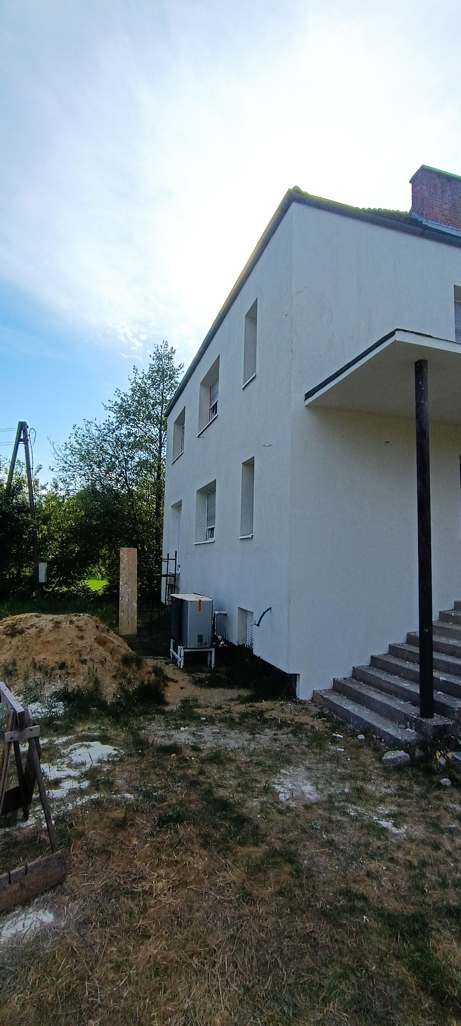 Dwupiętrowy dom z białą elewacją, widok z boku, z widocznym klimatyzatorem na zewnątrz, stertą piasku i drewnianym kozłem w trawie.