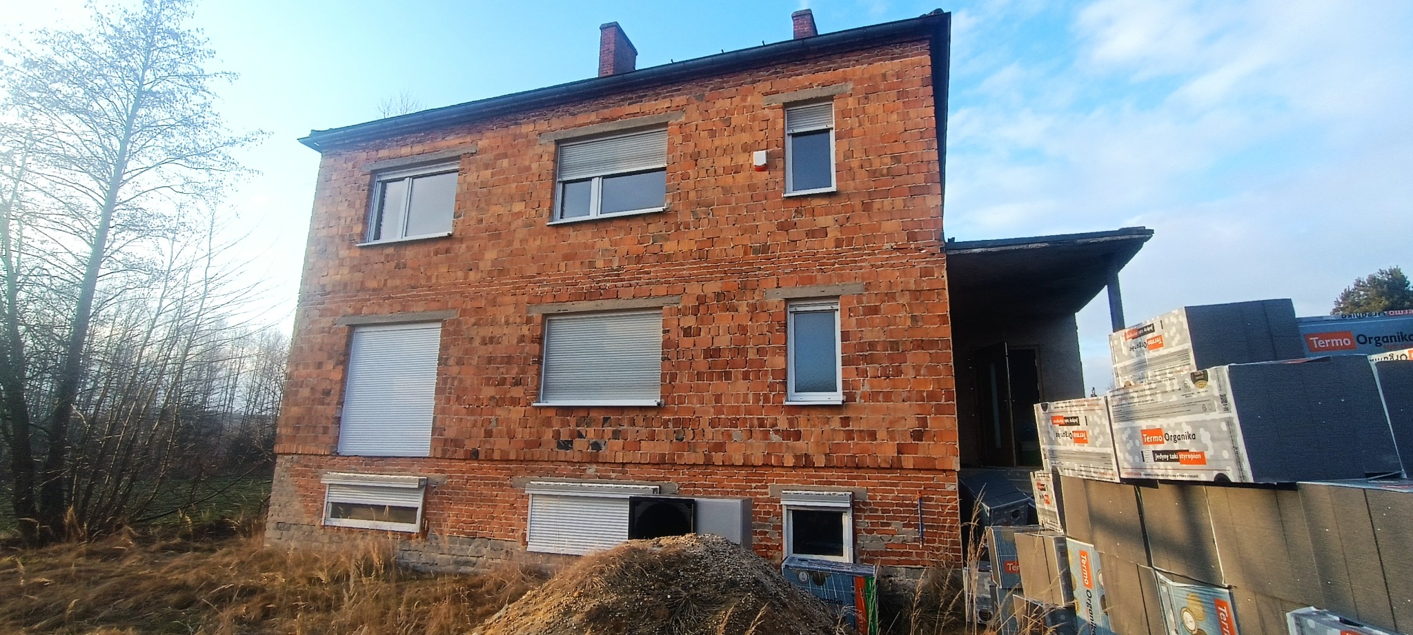 Dwupiętrowy budynek z czerwonej cegły z oknami z roletami, widoczne materiały izolacyjne Termo Organika, przygotowane do ocieplenia elewacji, zarośnięty teren wokół budynku.