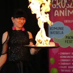 Kobieta w czarnej sukience z brokatem uśmiecha się, trzymając płonący rekwizyt na tacy podczas pokazu animacji dla dzieci na tle plakatu z ofertą usług eventowych.