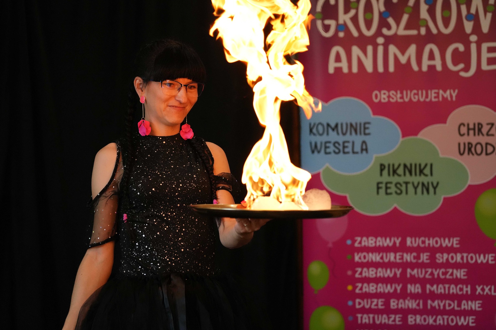 Kobieta w czarnej sukience z brokatem uśmiecha się, trzymając płonący rekwizyt na tacy podczas pokazu animacji dla dzieci na tle plakatu z ofertą usług eventowych.